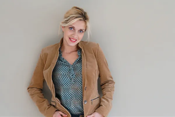 A smiling middle-aged woman in a gray top and tan leather jacket stands against a wall. Get Bioidentical Hormone Replacement Therapy services from Deborah Wilson, M.D. & Associates in Scottsdale.