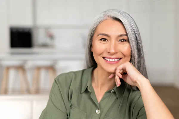 An asian woman with long gray hair and a green shirt sitting and smiling on her couch, happy with her menopause care from [NAME} in Scottsdale.